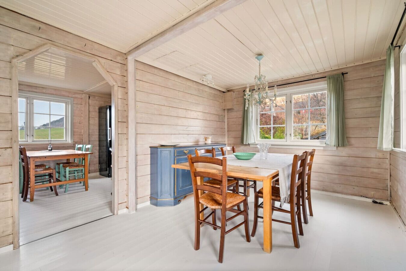 A dining area with a wooden table and chairs, a chandelier, and a view of the outdoors through a window.