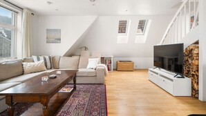 Una sala de estar moderna con sofá, mesa de centro y televisor. Se observa una escalera y una ventana con cortinas.