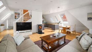 Amplia sala de estar con televisor, sofá y mesa de centro. Se ve una cocina al fondo y hay tragaluces.