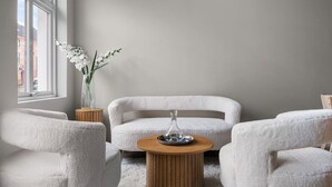 Un salon moderne avec une table basse ronde en bois, deux fauteuils blancs et un canapé blanc. Un vase avec des fleurs blanches est posé sur une table d’appoint, près d’une fenêtre.