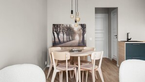 A dining area with a round table, four chairs, a painting of a tree-lined path, and a kitchen area with a sink and cabinets. - Bergen Beds Apartments | Urban Escape (Bergenhus)