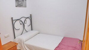 Une petite chambre avec un lit simple, dotée d’une tête de lit en métal, d’une table de chevet en bois, d’une couvre-lit blanche et d’une couverture rose.