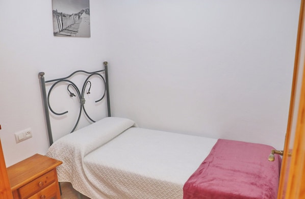 Una habitación pequeña con cama individual, cabecera de metal, mesita de noche de madera, colcha blanca y manta rosa.