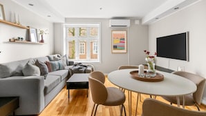 Ein modernes Wohnzimmer mit einem grauen Sofa, einem runden Esstisch mit Stühlen, einem Couchtisch und einem großen, schwarzen Fernseher an der Wand.