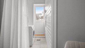 Un pasillo estrecho con una lavadora, una puerta que da al exterior y vistas a edificios por la ventana.