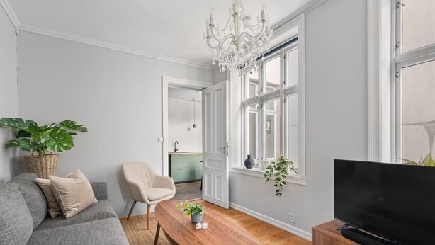 Una sala de estar moderna con un sofá gris, una mesa de centro de madera, un televisor sobre un mueble de madera, una araña y vistas a la cocina.