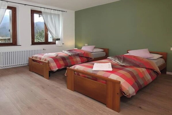 A room with two wooden beds, each with a plaid bedspread and pillows. There is a window with curtains, a radiator, and a view of greenery outside.