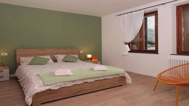 Un dormitorio con cama de madera, paredes verdes, una ventana con cortinas y una silla de mimbre.