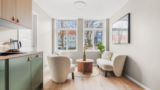 A modern living room with a wooden floor, a green cabinet, two white armchairs, a round wooden coffee table, and a view of buildings through large windows.