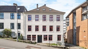 A multi-story building with a symmetrical facade, featuring a central entrance with a black door, multiple windows with red frames, and a flat roof with dormer windows.