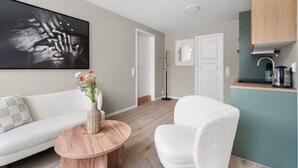 A modern living room with a sofa, a round wooden coffee table, and a white armchair. There is a painting on the wall and a kitchen area with a coffee machine.