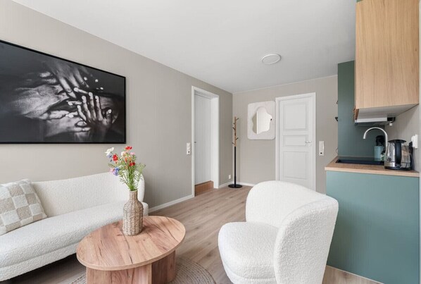 Una sala de estar moderna con un sofá, una mesa de centro redonda de madera y un sillón blanco. Hay un cuadro en la pared y una zona de cocina con una cafetera.