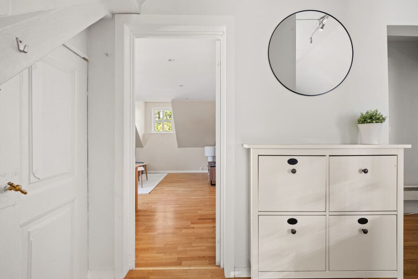 Un couloir avec une porte blanche, un plancher en bois, une armoire blanche avec des tiroirs et un miroir rond accroché au mur.
