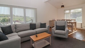 Uma sala de estar ampla com um sofá cinzento, poltronas e uma mesa de centro de madeira. Grandes janelas com persianas oferecem vista para o exterior.