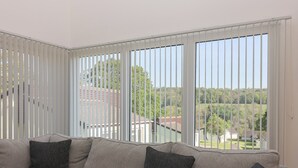 Uma sala de estar moderna com sofá em forma de L, janelas amplas com persianas e vista para uma paisagem verde.