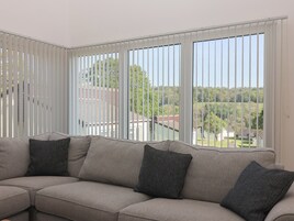 Una sala de estar moderna con un sofá modular, ventanas amplias con persianas y vistas a un paisaje verde.