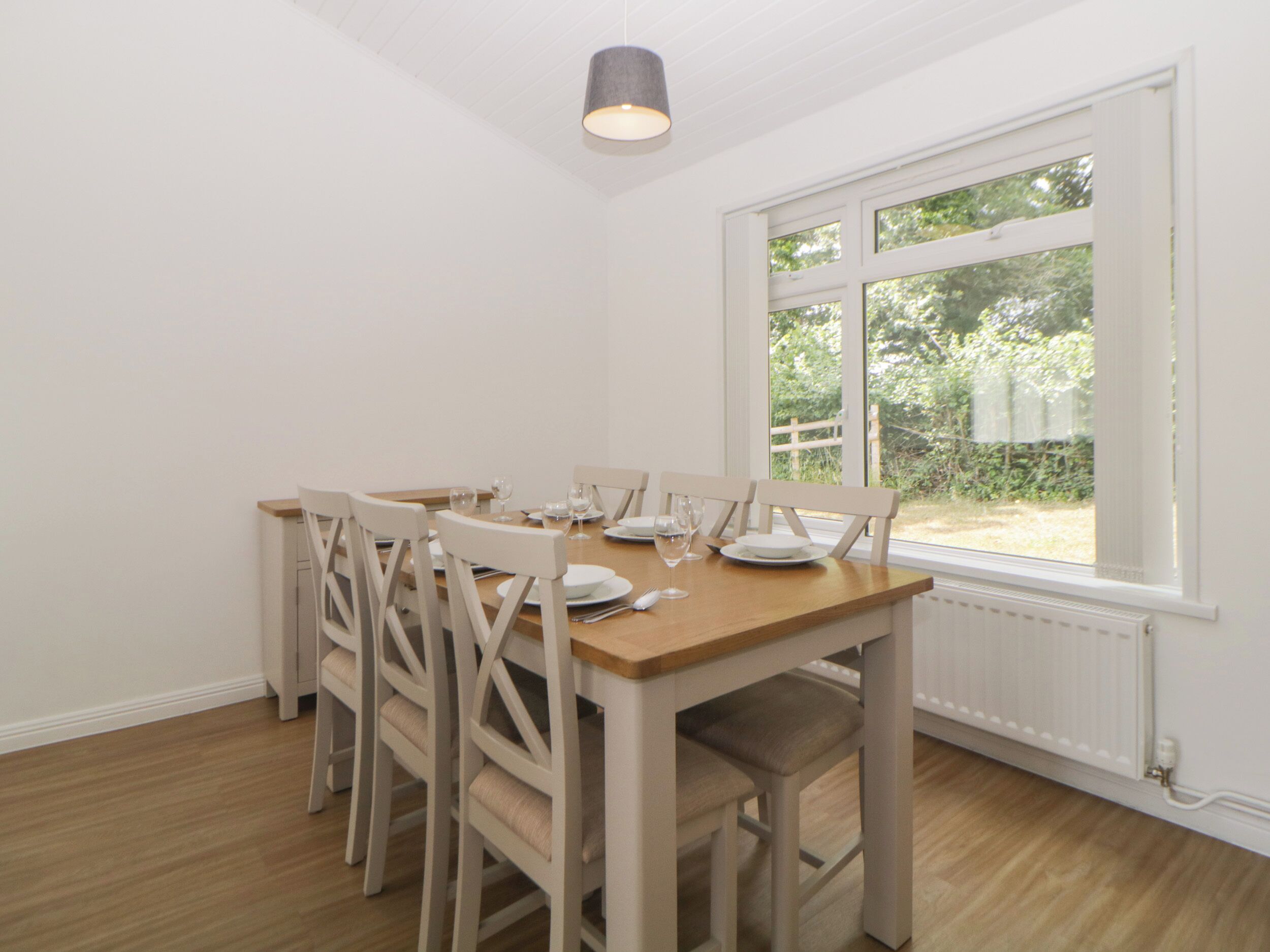 Un comedor con una mesa de madera y sillas blancas, una ventana con vistas a follaje y una lámpara en el techo.