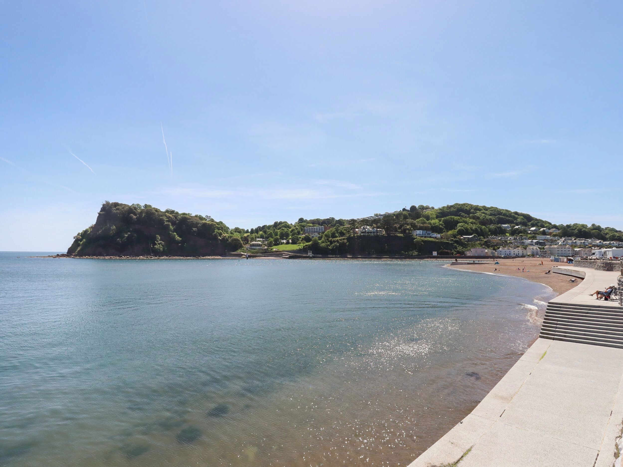 A coastal scene with a clear beach, a rocky outcrop, and buildings on a hillside.