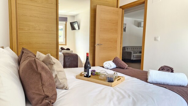 A hotel room with a large bed, wooden doors, a bedside table with a bottle and glasses, and a view into another room with a sofa and a TV.