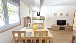 A modern kitchen with white cabinets and a black clock, a dining area with a wooden table and chairs, and a TV on a wooden stand.