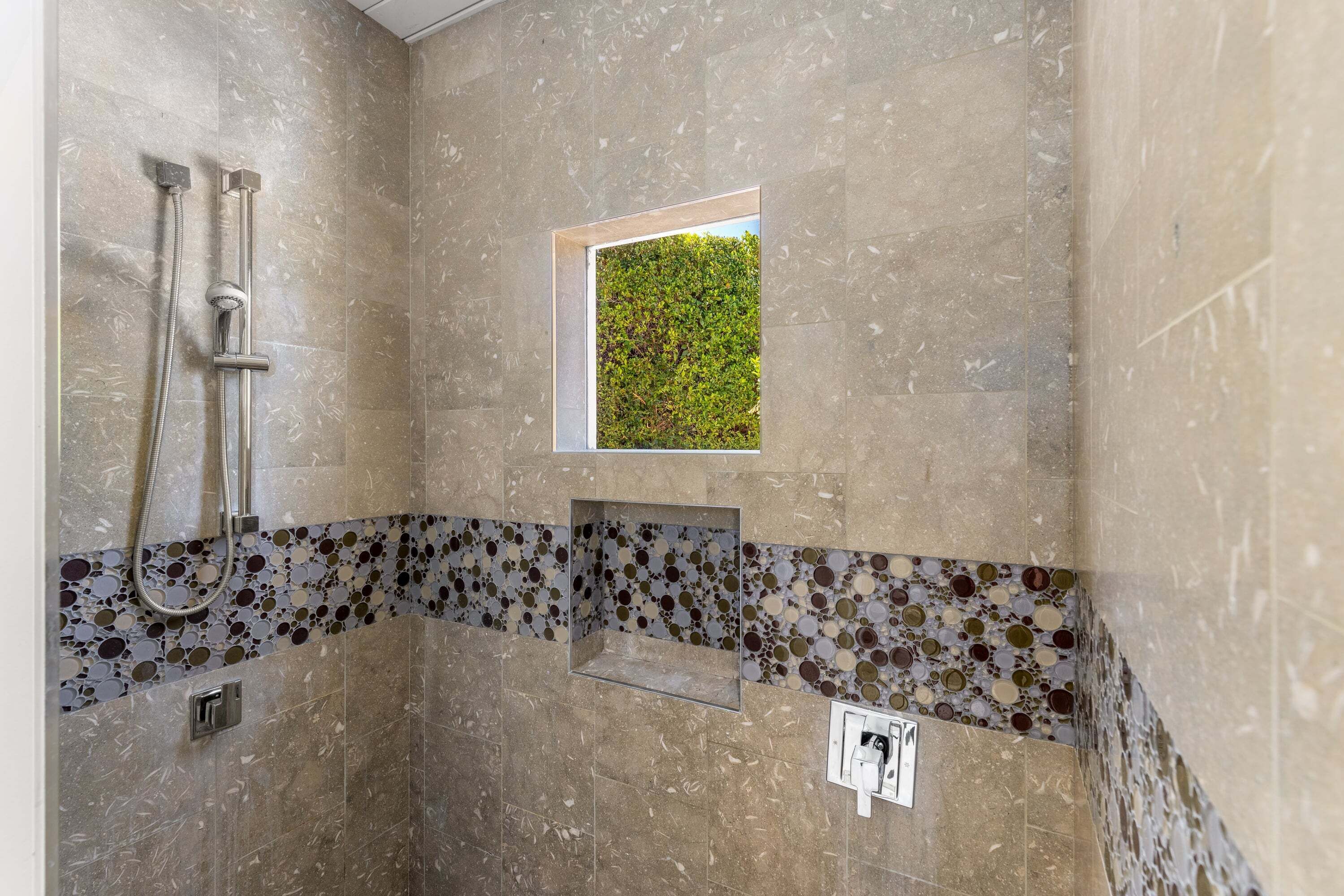 A shower with a handheld showerhead, a wall-mounted faucet, and a small window with a view of greenery.