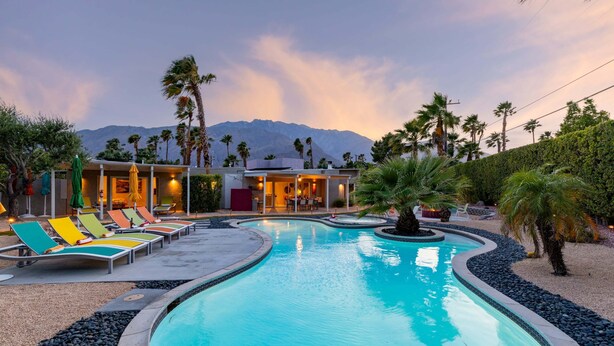 A swimming pool with lounge chairs, a building with multiple rooms, palm trees, and a mountain in the background.