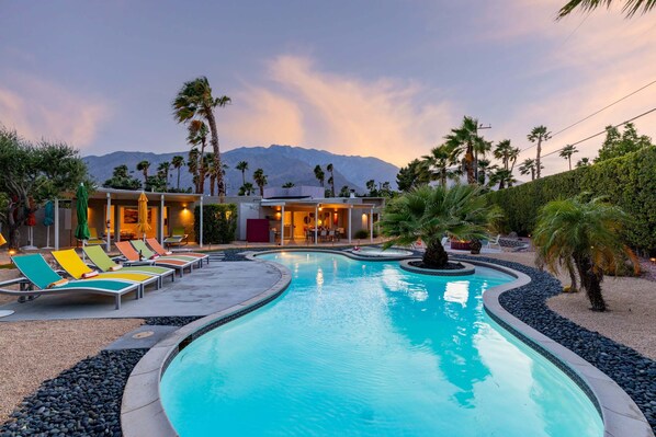 A swimming pool with lounge chairs, a building with multiple rooms, palm trees, and a mountain in the background.