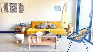 Un salon avec un canapé jaune, une table basse en bois, un fauteuil bleu et une planche de surf accrochée au mur.