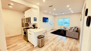 Una cocina moderna con una isla central, un comedor con alfombra negra y una sala de estar con sofá y sillones.