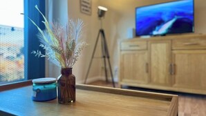 Une table en bois sur laquelle se trouve un vase rempli de fleurs séchées et un bocal. Un trépied et un téléviseur sont visibles à l’arrière-plan.