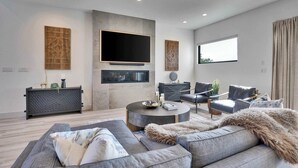 A modern living room with a large flat-screen TV, a fireplace, a sofa set, and a dining area with chairs.