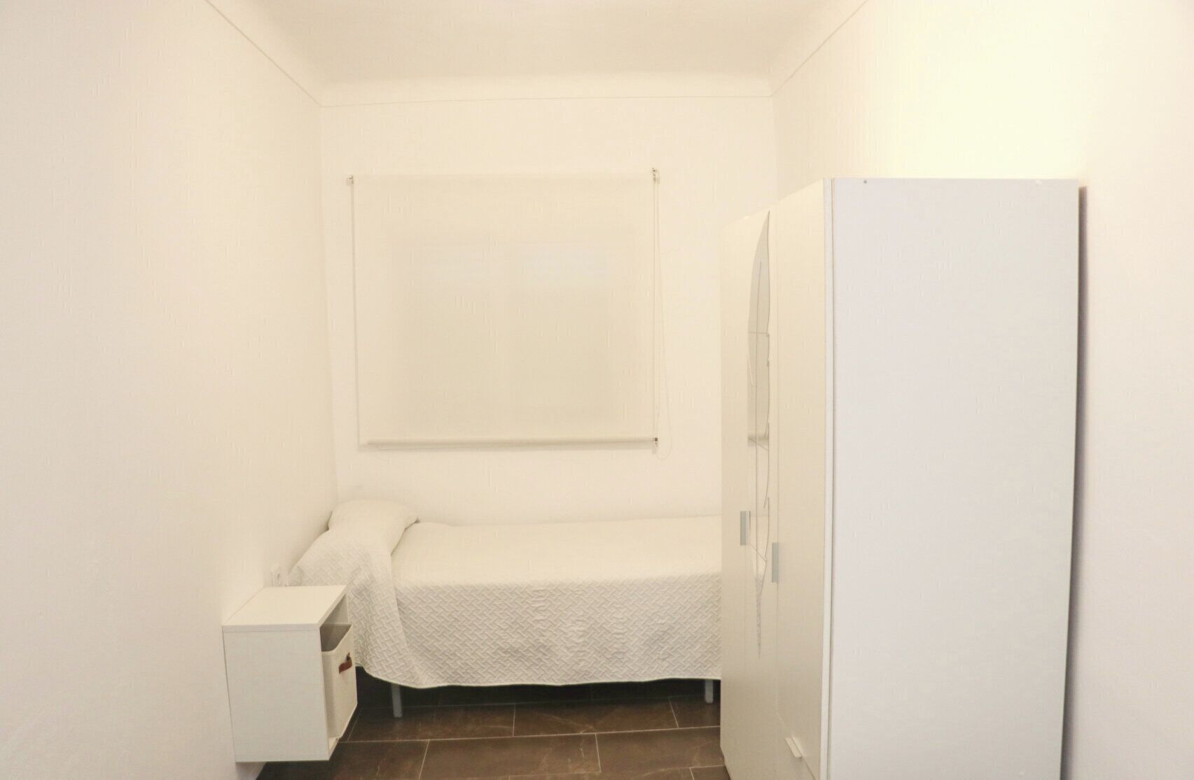 A small, white, single-bed room with a white wardrobe and a small bedside table.