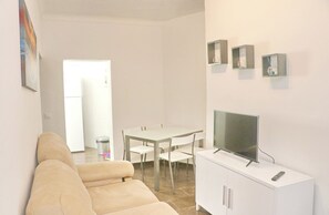 Un salon avec un canapé, un téléviseur posé sur un meuble, une table à manger avec des chaises, et un coin cuisine à l’arrière-plan.