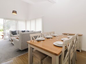 Un comedor con una mesa de madera dispuesta para seis comensales, sillas blancas y un sofá gris al fondo.