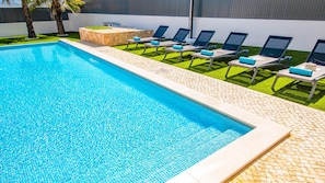 A rectangular swimming pool with clear blue water, surrounded by a stone border. There are several lounge chairs with blue cushions aligned along the pool's edge.