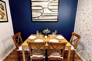 A dining table set for four with wooden chairs, a blue wall, and a framed abstract painting above.