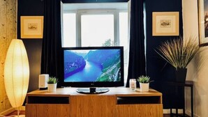 Un soggiorno moderno con un mobile TV in legno, una TV a schermo piatto che mostra un paesaggio fluviale, una lampada da terra e due piante in vaso.
