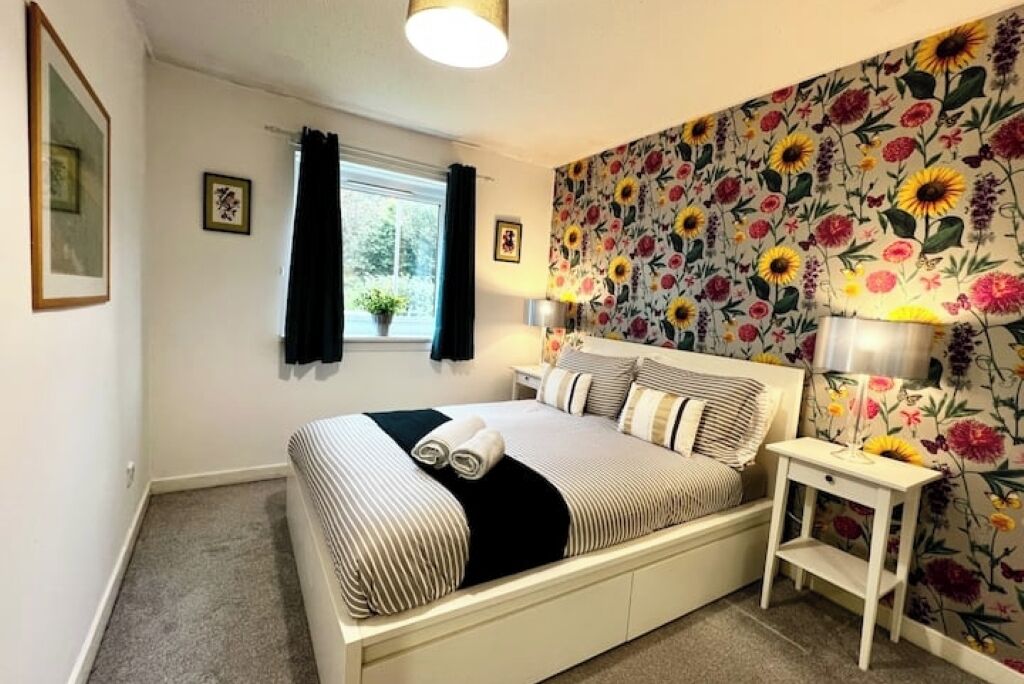 A neatly made bed with striped bedding, a small bedside table with a lamp, and a floral wallpaper.