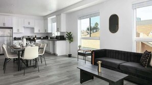 A modern kitchen with white cabinets, a dining area with a table and chairs, and a black sofa in a living area with large windows. - Urban 3BR Retreat with Modern Amenities (Los Angeles)