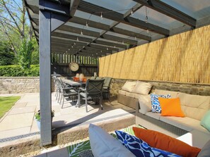 En udendørs patio med et overdækket siddeområde, stenvæg og et ur på væggen.