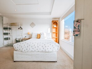 Un dormitorio con una cama grande, una estantería de madera, un escritorio con una lámpara y una ventana con vistas a edificios.