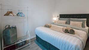 Quarto com cama, um cabideiro, mesa de cabeceira com luminária e prateleira na parede.