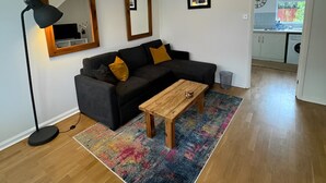 Un salon moderne avec un canapé noir, une table basse en bois et un tapis coloré. On y trouve deux lampes fixées au mur et un grand miroir au-dessus du canapé.