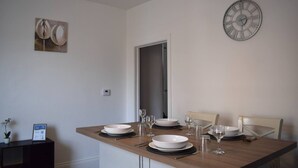 A dining area with a wooden table set for four, including plates, bowls, glasses, and cutlery. A wall-mounted clock is visible above the table. - The Twickenham Apartment Near Strawberry Hill (Twickenham)