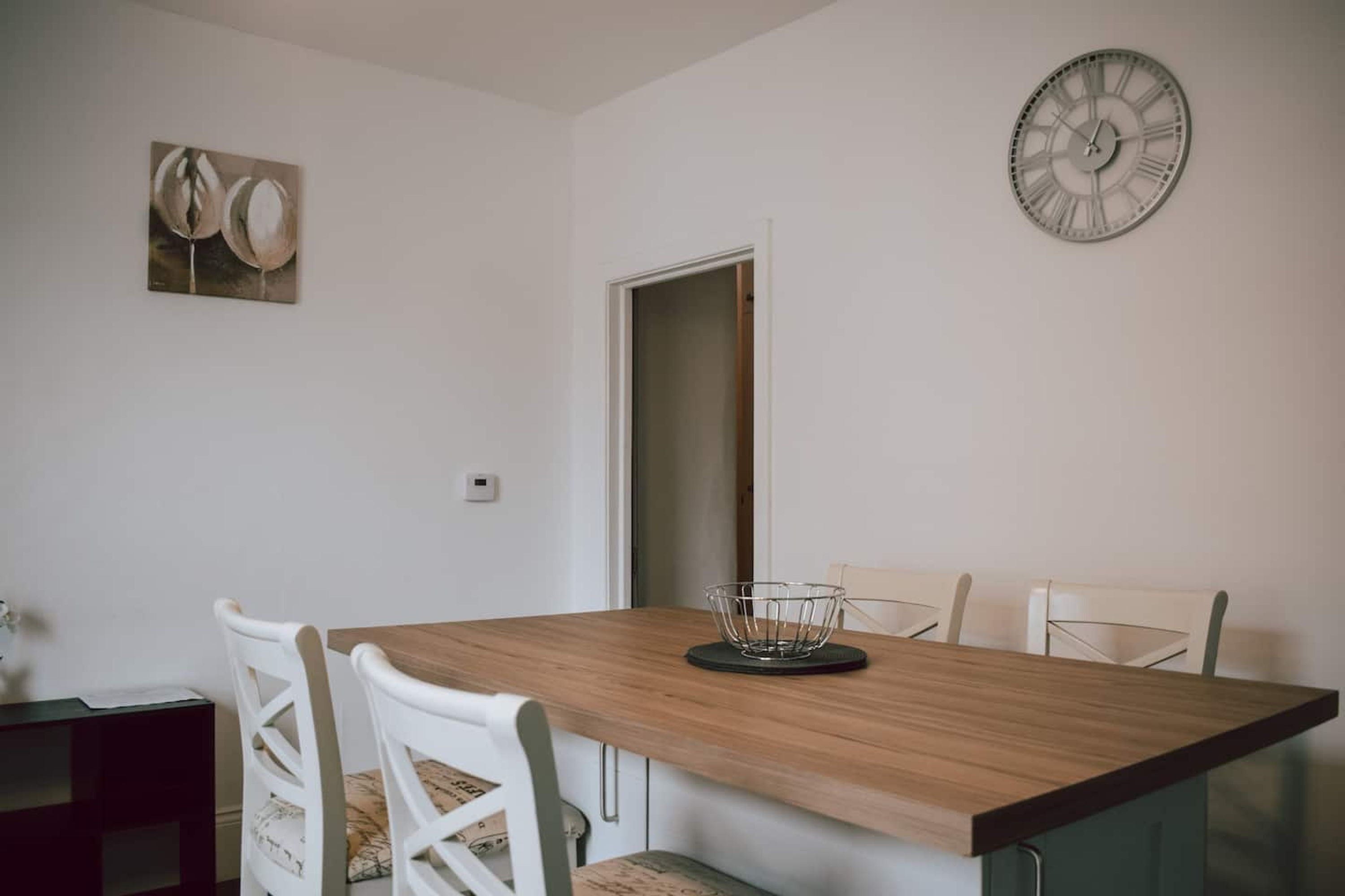 A dining area with a wooden table, white chairs, a clock, and a door.