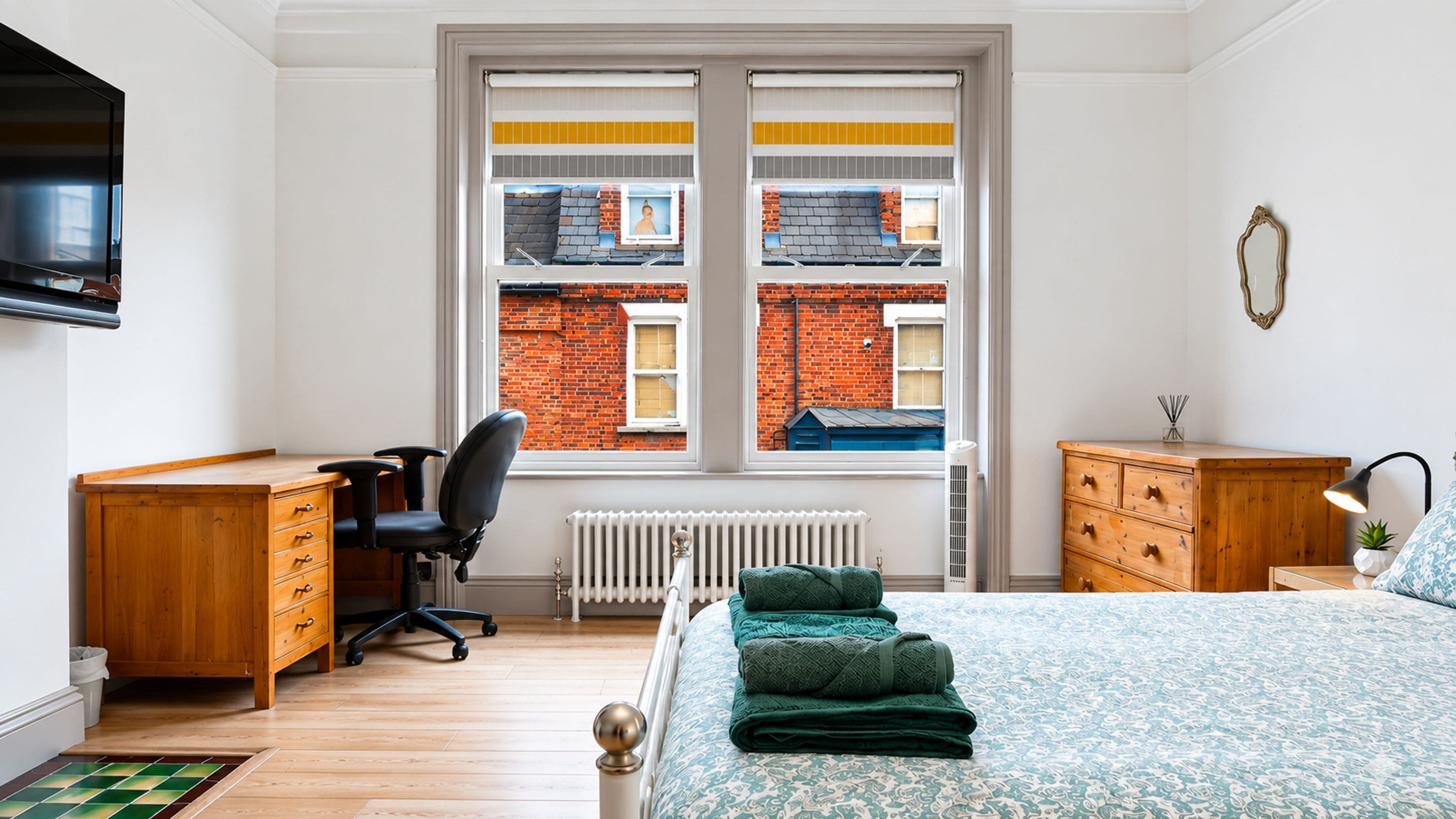 A bedroom with a bed, a desk, a chair, a radiator, and a window with a view of a brick building.