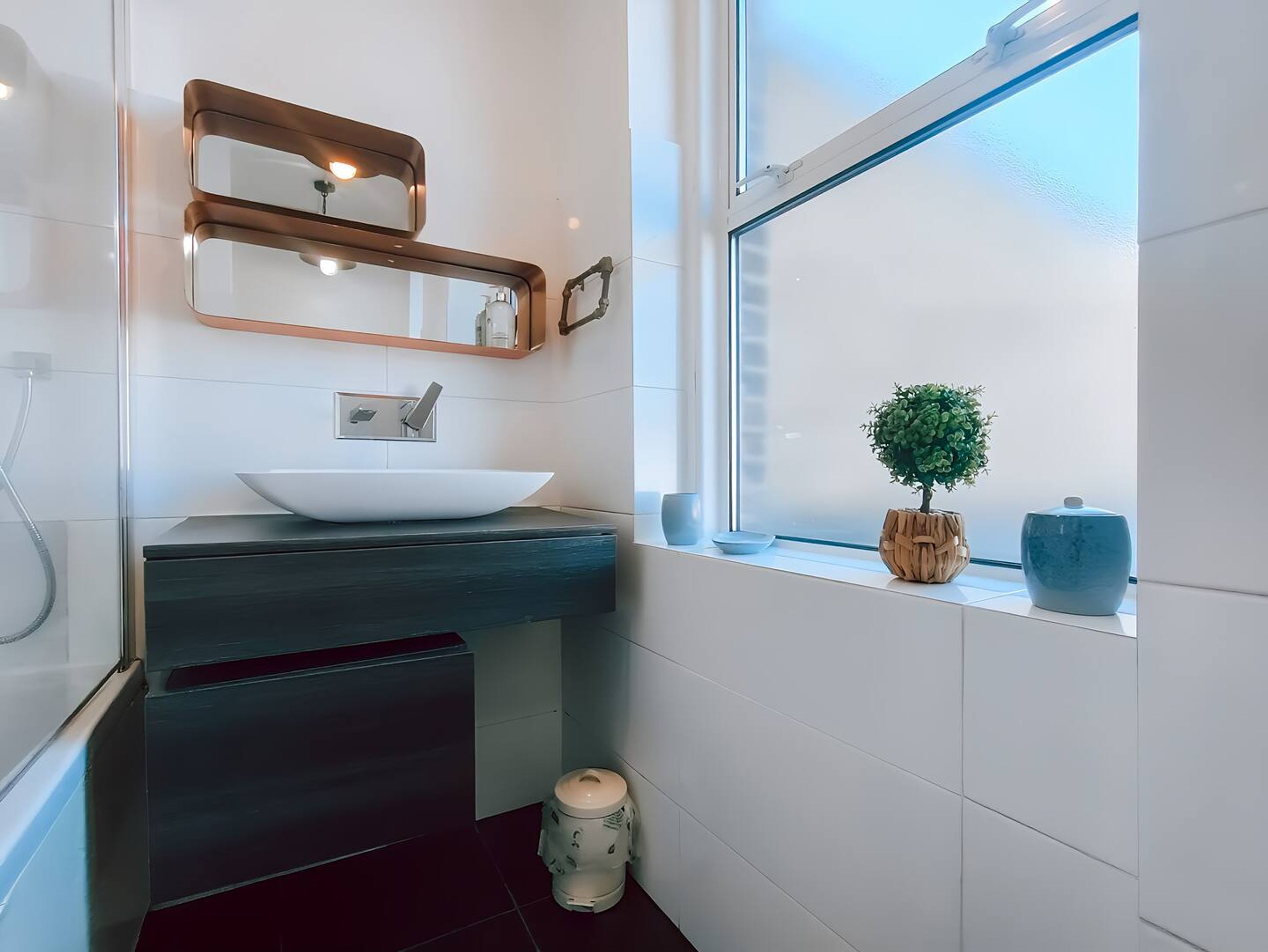 A modern bathroom with a white sink, a mirror, and a window with a view of a tree and a blue vase.