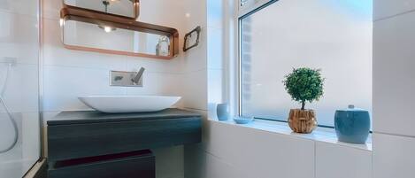 A modern bathroom with a white sink, a mirror, and a window with a view of a tree and a blue vase.