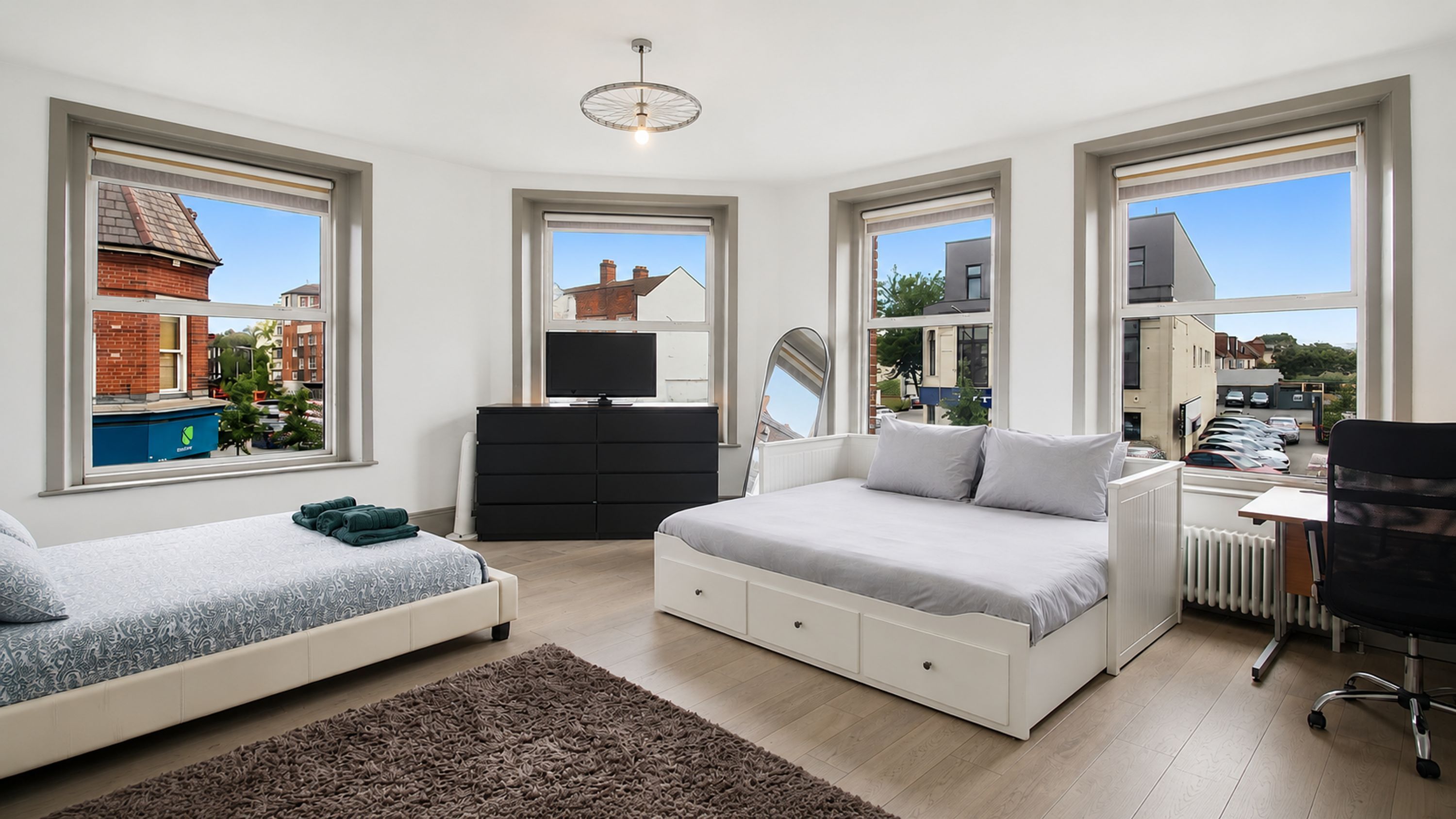 A modern bedroom with two beds, a dresser, a desk, and large windows offering views of the outside.