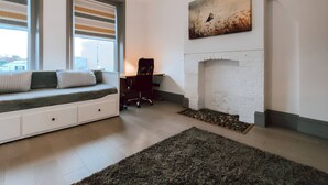 A modern living room with a white brick fireplace, a large window with blinds, a black office chair, and a grey rug. - The Twickenham Apartment Near Strawberry Hill (Twickenham)